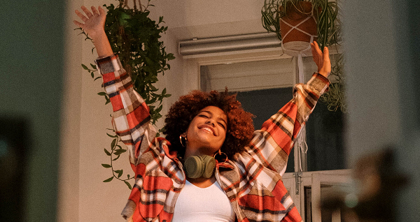 Une jeune femme joyeuse danse chez elle, les bras levés en l'air, entourée de plantes suspendues. Elle porte un casque autour du cou et une chemise à carreaux sur un t-shirt blanc.